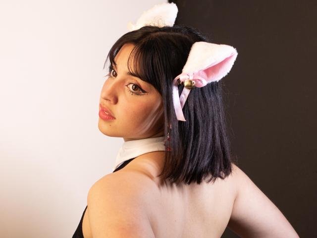 VickyButerfly with short black hair, wearing white cat ears with pink bows, looking over shoulder, black top, white neck ribbon, light skin, pink lips, subtle makeup, black background.