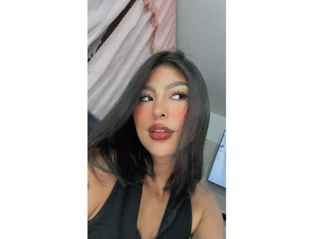 Carla Russo, com cabelo preto na altura dos ombros, blush vermelho e lábios vermelhos brilhantes, vestindo um top preto de alças, olhando para o lado com um leve franzido..