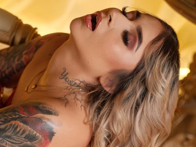 AbbyAmazing with her eyes closed, mouth open, head tilted back, showing tattoos on her neck and arm, blonde hair, and a gold necklace. Her tits are partially visible.