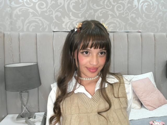 MelodySwet with brown pigtails, wearing a beige sweater, white shirt, and choker, has small tits and a cute face, sitting on a bed with a gray lamp.