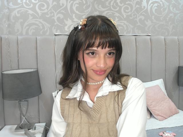 MelodySwet has brown hair with flowers, wearing a beige sweater over a white shirt, pearl necklace, and smiling. Her tits are small and she's sitting on a bed.