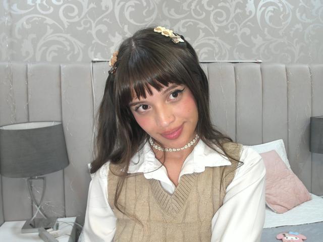 MelodySwet has brown hair, bangs, and small floral hair clips. She's wearing a beige sweater over a white shirt, with a pearl necklace, smiling at the camera.