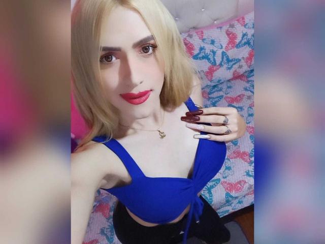 LorenaFoxy with blonde hair, blue crop top, red lipstick, long nails, and a ring, showing her tits, sitting on a pink and blue floral-patterned bed.