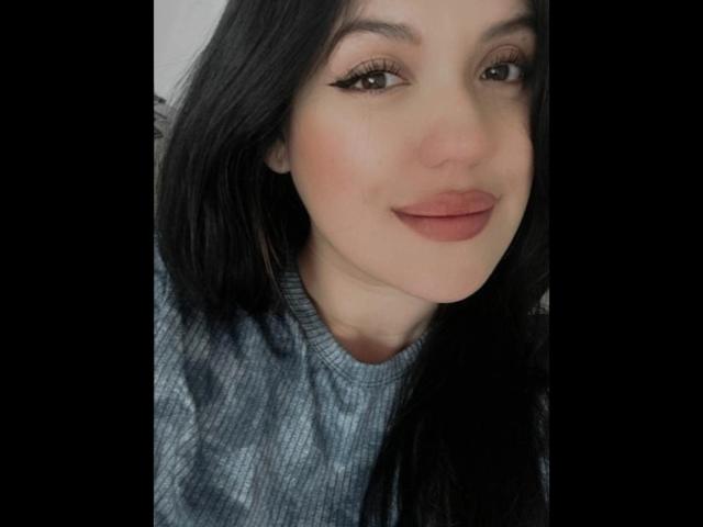 LauraRichis with black hair, light skin, and subtle makeup, wearing a gray sweater, smiling with pink lipstick, and small, pointed eyeliner. Her tits are not visible.