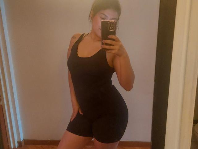 LauraRichis in a black tank top and shorts, taking a mirror selfie, showing her curvy ass and small tits, with one hand on her hip, in a dimly lit room.
