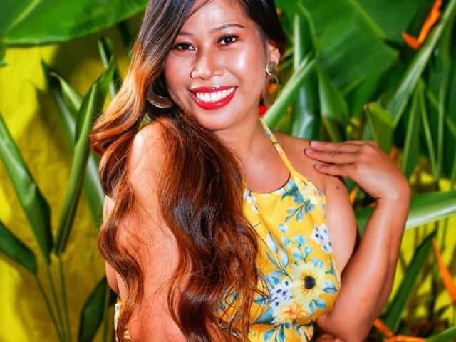 FilipinaBeauty69 com cabelos castanhos longos e ondulados, vestido floral amarelo, batom vermelho, sorrindo, mão perto do rosto, fundo com folhagem verde, usando brincos de argola, com uma aparência fofa e sexy..