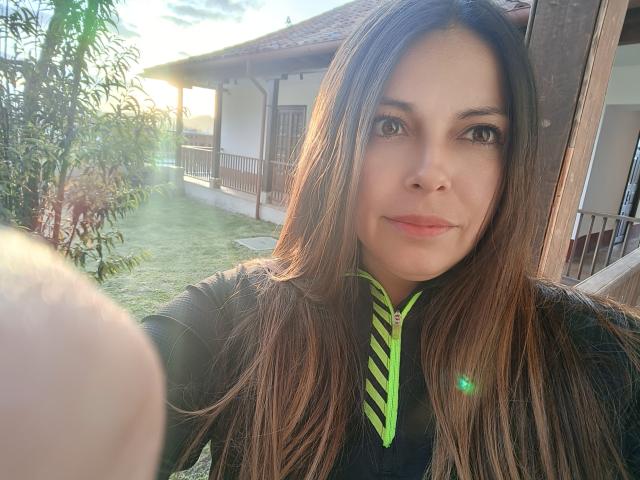 Seher a de longs cheveux bruns, une chemise rayée de vert, une veste noire et un léger sourire. On peut voir ses yeux bruns et sa peau claire. Une maison baignée de soleil et une pelouse verte se trouvent en arrière-plan..