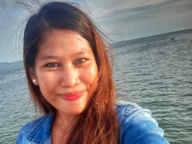 FilipinaBeauty69 con lunghi capelli castani, un sorriso carino, rossetto rosso, piccoli orecchini e una giacca di jeans, in piedi vicino a un oceano calmo, con il viso illuminato dalla dolce luce del tramonto..