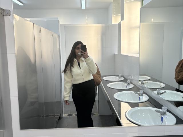 DianaFernandezz with long brown hair, white zip-up jacket, black pants, standing in a bathroom, taking a mirror selfie, showing her ass, in front of multiple sinks.