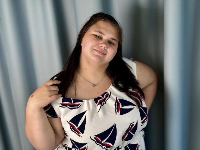 KaidoKun tiene senos grandes, piel clara y cabello castaño. Lleva un vestido blanco con estampados de velas azules y rojos, y un collar de plata. Está sonriendo suavemente..