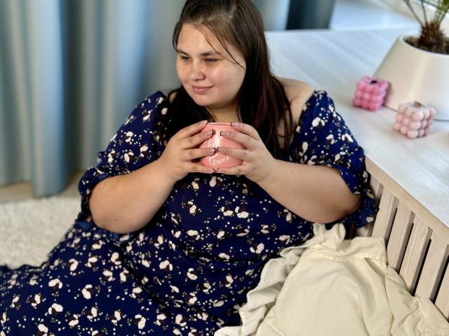 KaidoKun, con un seno di media grandezza, indossa un abito blu con fiori, tiene una tazza rosa, è seduto su un letto bianco, sorride leggermente, ha i capelli castani, la pelle chiara, e l'ambientazione è un contesto domestico informale..