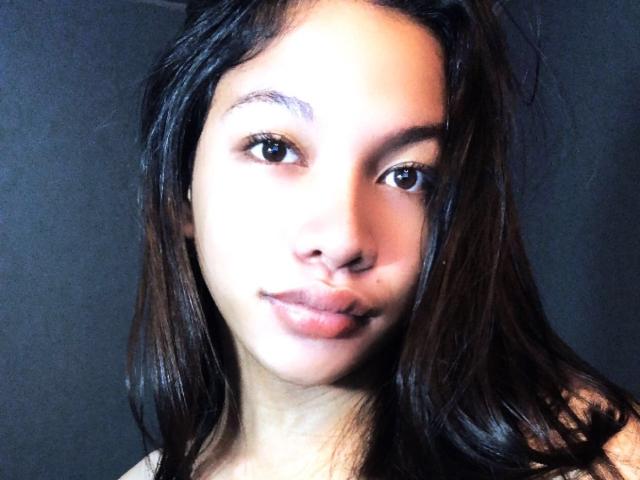 StaceGoddess has light brown skin, dark brown eyes, and long black hair, with full lips slightly parted in a subtle smile. Her face is centered and lit from the front.