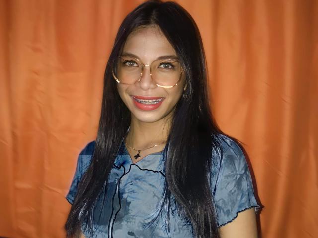 StaceGoddess with long black hair, wearing glasses, blue tie-dye shirt, smiling, orange curtain background, small black necklace with heart pendant.