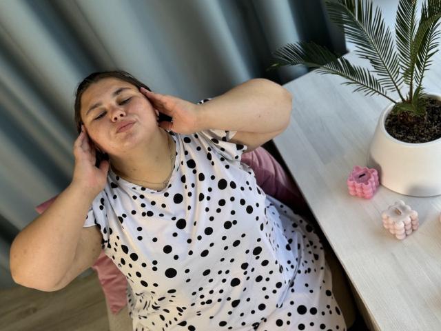 KaidoKun, avec un visage rond, des cheveux bruns, portant une robe blanche à pois, les mains potelées sur les oreilles, assis sur une chaise rose, une table avec une plante et un élastique rose..