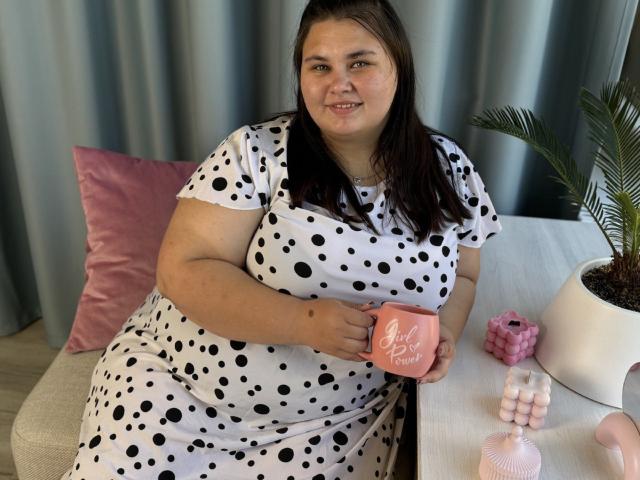 KaidoKun, a chubby woman with long brown hair, wears a white dress with black polka dots, holding a pink "Sweet Treat" mug, sitting near a pink pillow and plant.