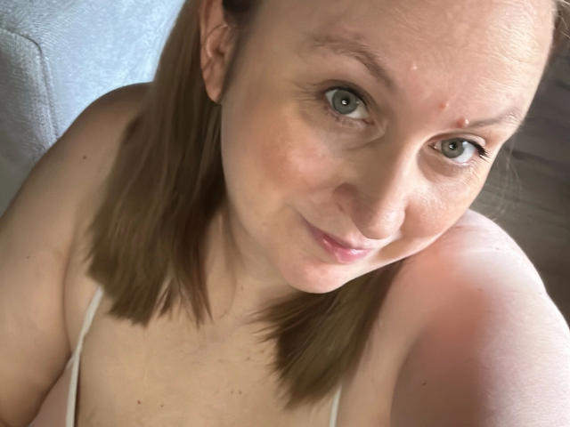 Noralina-hot with light brown hair, green eyes, and a subtle smile, showing her tits in a white strap top, looking up at the camera. Her skin is slightly pink.