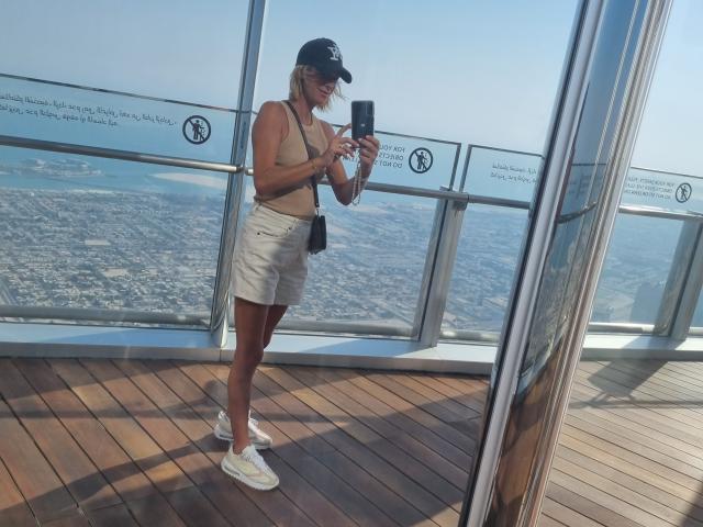 BlackAngeel with blonde hair, wearing a black cap, beige top, white shorts, and white sneakers, standing on a wooden deck, taking a selfie, showing her tits.