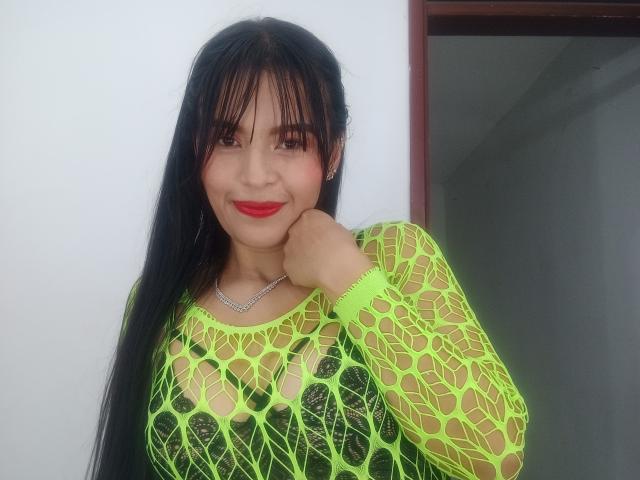 SusanKartier with long black hair, neon green fishnet top showing her big boobs, red lipstick, and a confident smile, resting her chin on her hand.