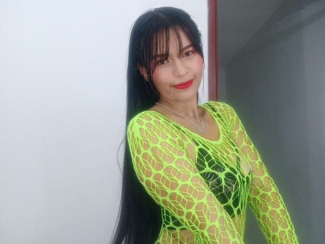 SusanKartier with long black hair, wearing a neon green fishnet top, showing her tits through the net, smiling with red lipstick, standing against a white wall.