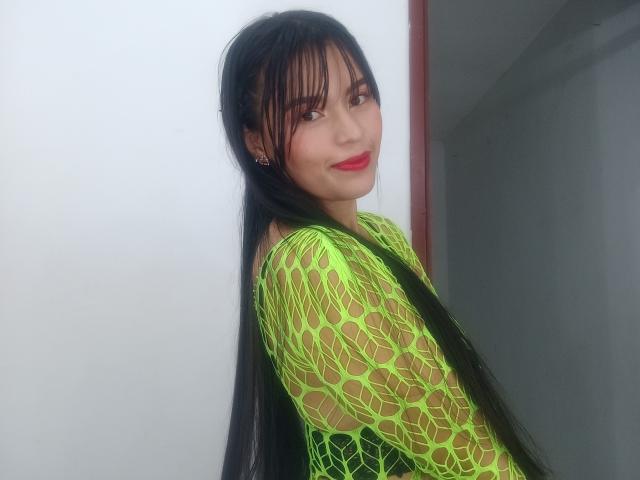 SusanKartier with long black hair, green fishnet top, red lipstick, smiling, looking over shoulder, showing her tits through the mesh, standing against a white wall.