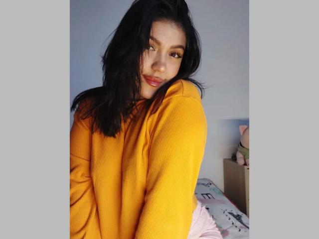 LauraRichis with black hair, yellow sweater, and pink pants, smiling slightly, looking over shoulder, soft light, casual, cute, sexy, medium tits, subtle makeup, relaxed bedroom setting.