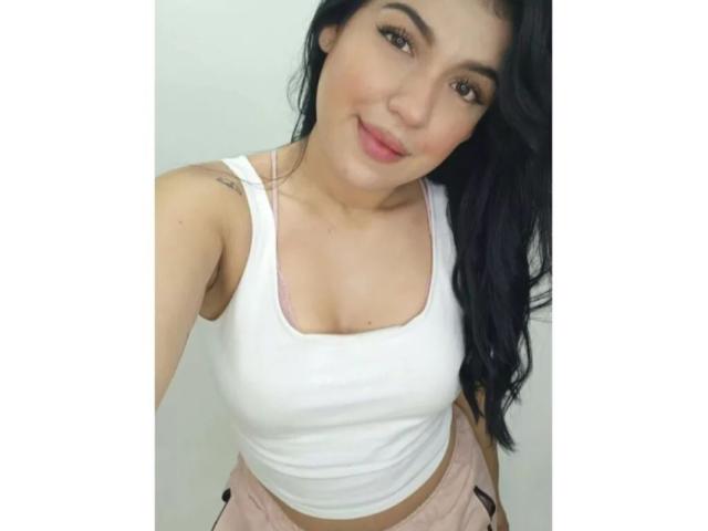 LauraRichis has black hair, brown eyes, and is wearing a white tank top showing her small tits, with pink shorts, smiling at the camera.