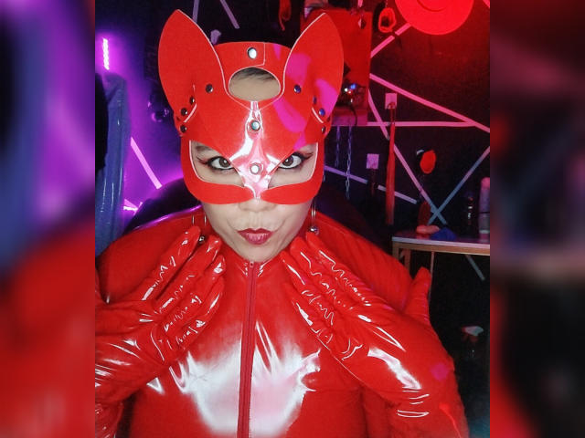 LexyDom wearing a shiny red latex cat mask with ears, red latex suit, and red lipstick, looking directly at the camera with a serious expression. Purple and red lights.