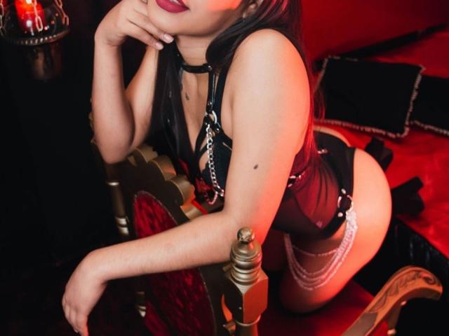 AmyXPlata, con cabello negro largo, lápiz labial rojo, arnés de cuero negro y ligas, arrodillada sobre una silla de terciopelo rojo, con la mano cerca de la boca, mostrando su trasero y un pequeño lunar..