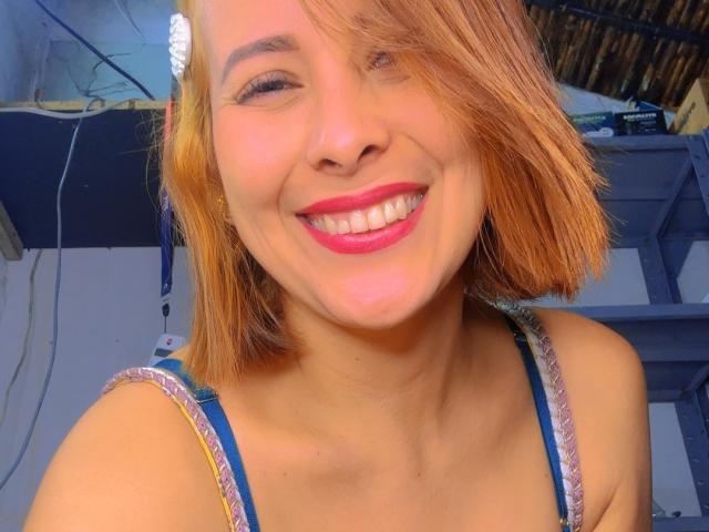 Sarita Velez, com um rosto sorridente, batom rosa e um corte de cabelo curto laranja, vestindo uma blusa listrada em azul e rosa, mostrando seus dentes e um sorriso levemente ondulado..