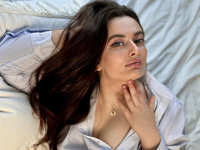 MilanaPrice with long dark brown hair, wearing a white patterned shirt, has her hand on her neck, looking at the camera, showing her tits slightly visible, on a white bed.