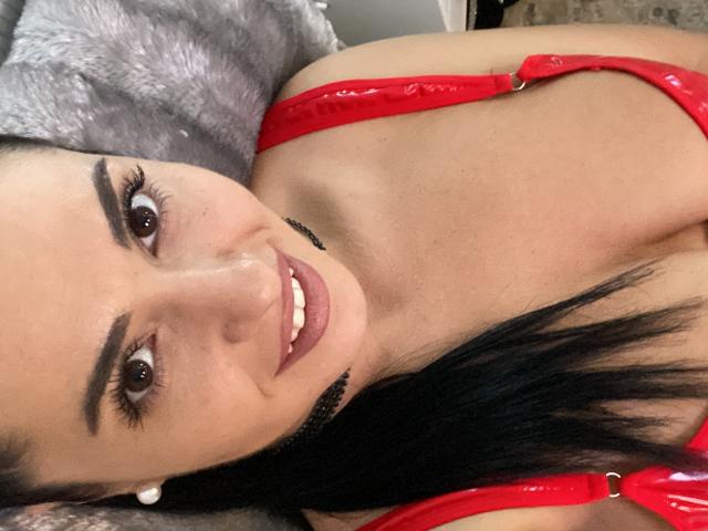 Jenniferwild69 with red latex bra, smiling at camera, brown eyes, black eyebrows, long black hair, pink lips, pale skin, lying on gray pillow, wearing pearl earrings.
