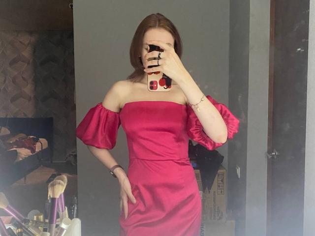 KiraNightly in a red off-shoulder dress with puffy sleeves, holding a phone in front of her face, standing with one hand on her hip, showing her tits.