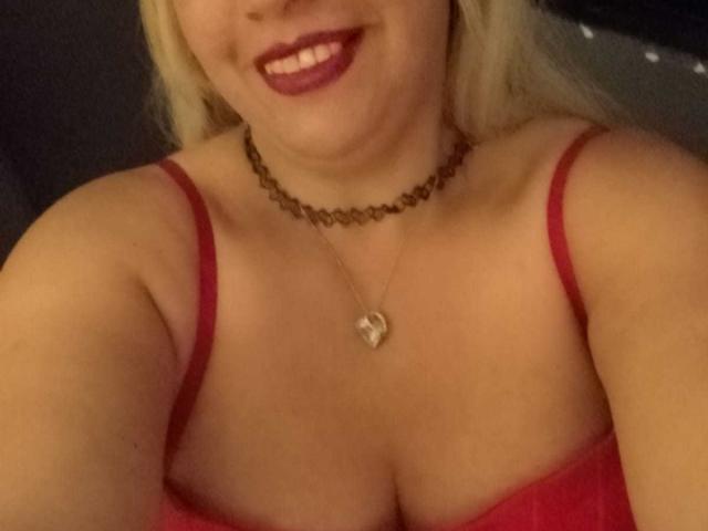 NathaLys with blonde hair, red dress, and necklace, smiling with dark red lipstick, showing cleavage, and a heart pendant. Boobs visible. Soft lighting.