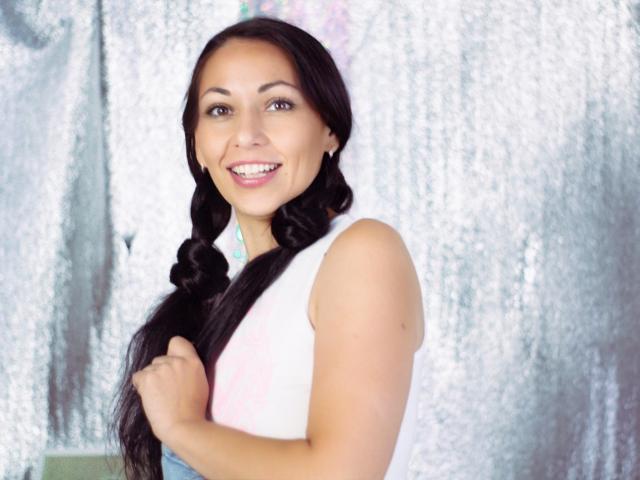 LiluGrants with long black braids, smiling, holding her tits, wearing a white sleeveless top, standing against a shiny silver backdrop, looking cute and sexy.