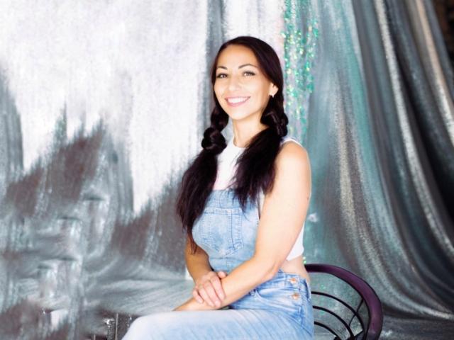LiluGrants with long black pigtails, wearing a white crop top and blue denim overalls, smiling, arms crossed, sitting on a black chair, showing her ass.