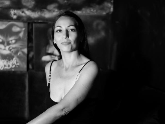 Foto em preto e branco de LiluGrants, com seios médios, vestindo uma blusa com alças finas, sentada em um ambiente escuro e melancólico, sorrindo suavemente, com cabelos castanhos longos, pele clara e aparência elegante..