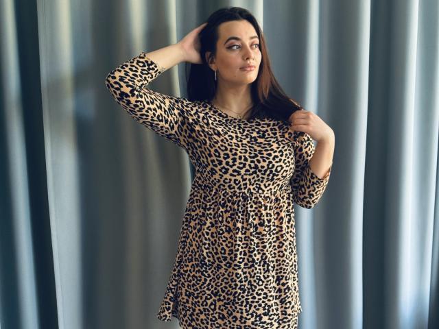 MilanaPrice with dark hair, wearing a leopard print dress, standing with hand in hair, looking slightly to the side, showing her ass and tits in a casual pose.