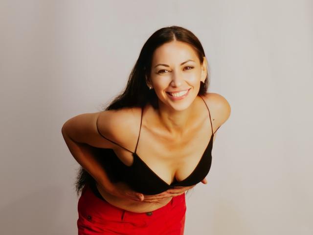 LiluGrants is smiling, leaning forward, hands on her tits, wearing a black bra, red pants, and long brown hair, with a cute, sexy look.