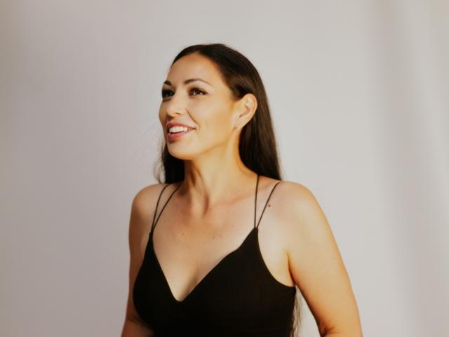 LiluGrants, smiling woman with long dark hair, wearing a black spaghetti-strap top, showing her tits, standing against a white wall, looking to the side.