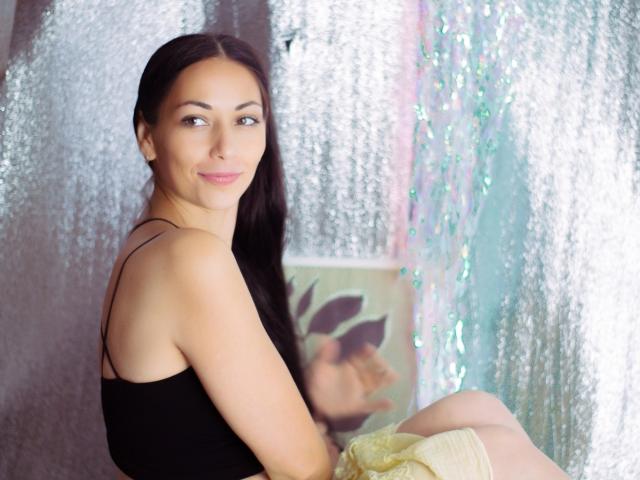 LiluGrants with long dark hair, black tank top, and subtle smile, looking over shoulder, sitting with yellow fabric, showing smooth ass, and light skin, in a shiny textured background.
