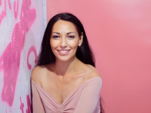 LiluGrants tiene el pelo largo y castaño oscuro, una sonrisa encantadora, y lleva una blusa rosa con un hombro descubierto, que deja ver sus pequeños senos. Está de pie frente a una pared rosa..