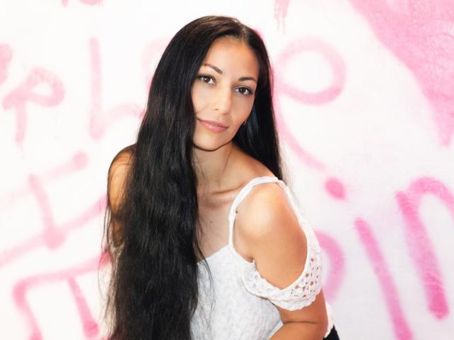 LiluGrants with long black hair, wearing a white off-shoulder lace top, smiling softly at the camera. She has cute tits and a slim, attractive face.