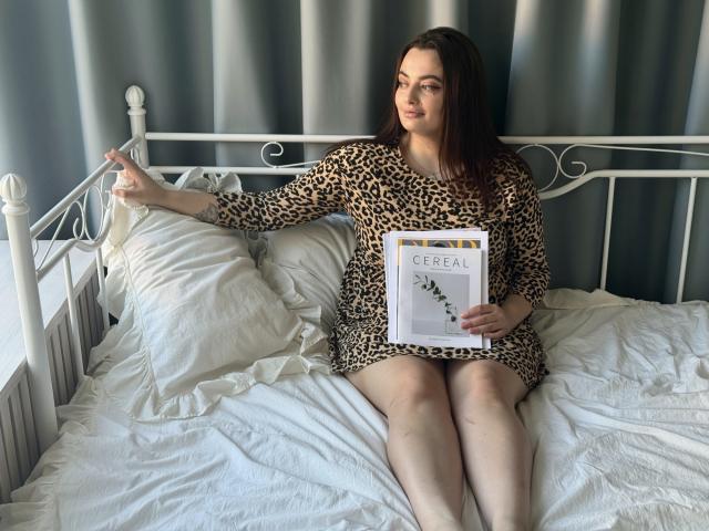 MilanaPrice, con la pelle chiara, i capelli castano scuro, indossa un vestito con stampa leopardo, è seduta su un letto bianco, tiene un libro in mano, indica con la mano destra, le gambe sono unite, ha un'espressione rilassata..