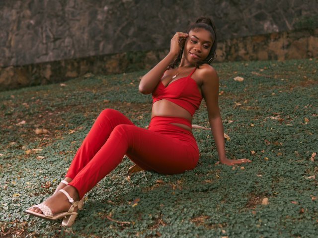 AlanaPatel, sitzend auf grünem Gras, trägt ein rotes Crop-Top und eine Hose, wobei ihre Brüste, ihr Hintern und ihre braune Haut sichtbar sind. Sie trägt weiße Sandalen, hat eine Hand im Haar und lächelt..