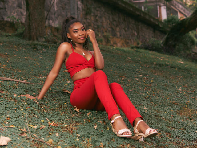 Alana Patel, mit dunkler Haut, kleinen Brüsten und einem hübschen Hintern, sitzt auf dem Gras, trägt ein rotes Crop-Top und eine Hose, berührt ihr Ohr, trägt weiße Sandalen und befindet sich in einem Wald..