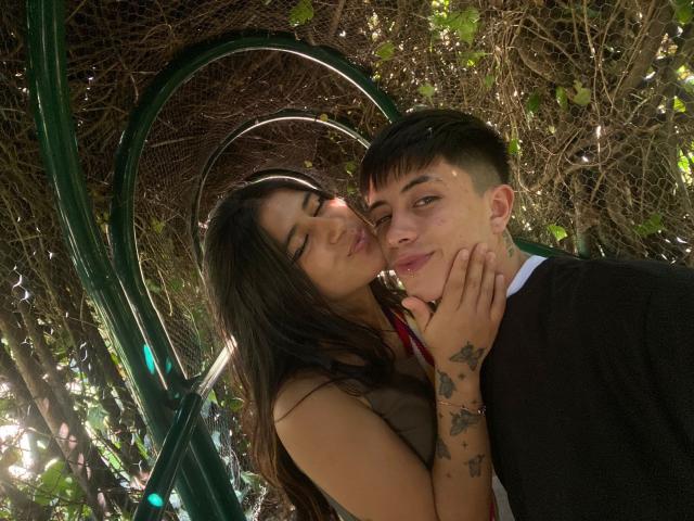LucyAndPerceo with long dark hair, kissing cheek, hand on Perceo's face, tattoos on hand, brown sleeveless top, black shirt, short dark hair, green metal structure, leafy background.