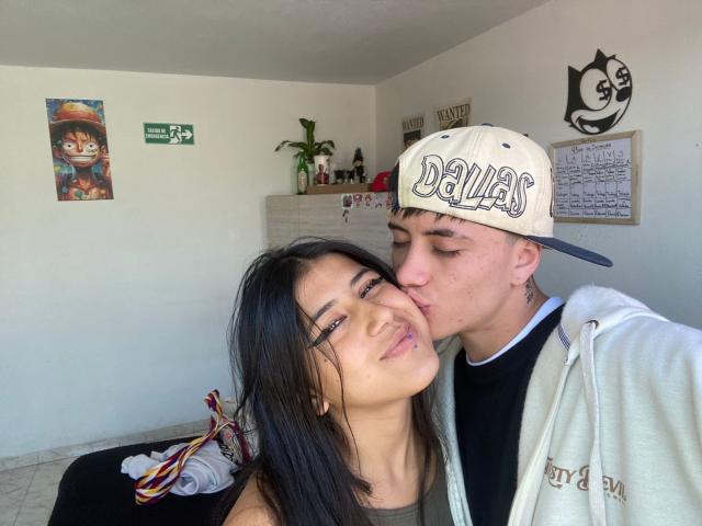 LucyAndPerceo, with black hair and tan skin, has her lips slightly parted as Perceo, with a white "Dallas" cap, kisses her cheek. She's wearing a green top, he's in a hoodie.