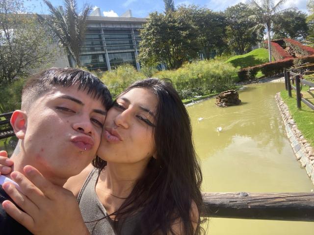 LucyAndPerceo, a chubby dude with short black hair and small red pimple, kisses LucyAndPerceo's brown-haired, tan-skinned face with pink lipstick, by a green pond.