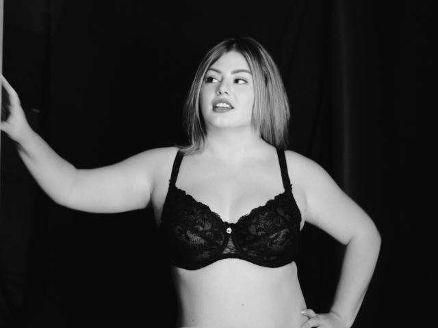 ZannaDreil with medium tits, wearing a black lace bra, has light skin, blonde hair, and a slightly open mouth, standing with one arm raised against a dark background.