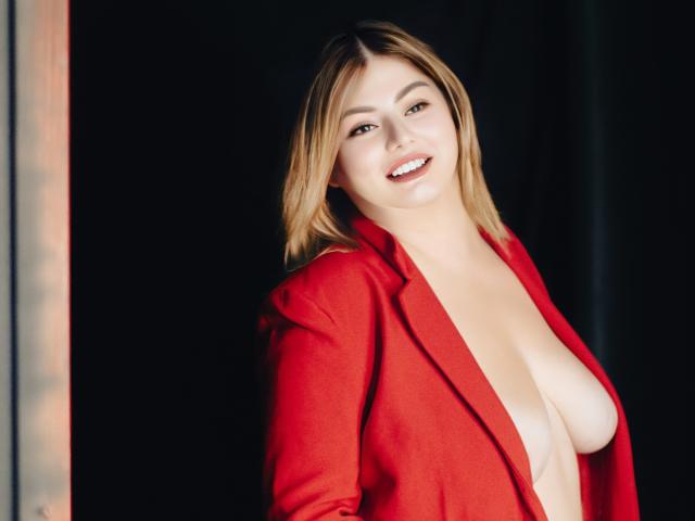 ZannaDreil with blonde hair, smiling, wearing a red blazer, exposing her big tits, standing in front of a dark background, looking at the camera.